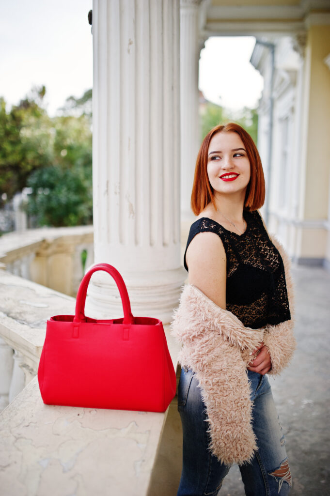 Fille aux cheveux roux avec un sac à main rouge posée près d'une maison ancienne.